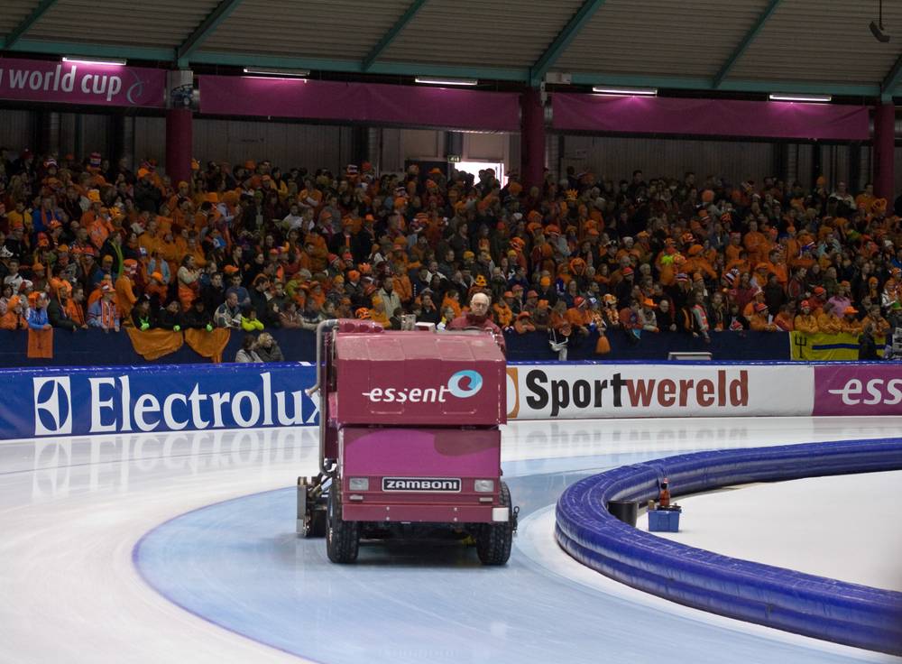 A Zamboni ice resurfacer circles the speed skating oval as an orange-clad crowd watches.