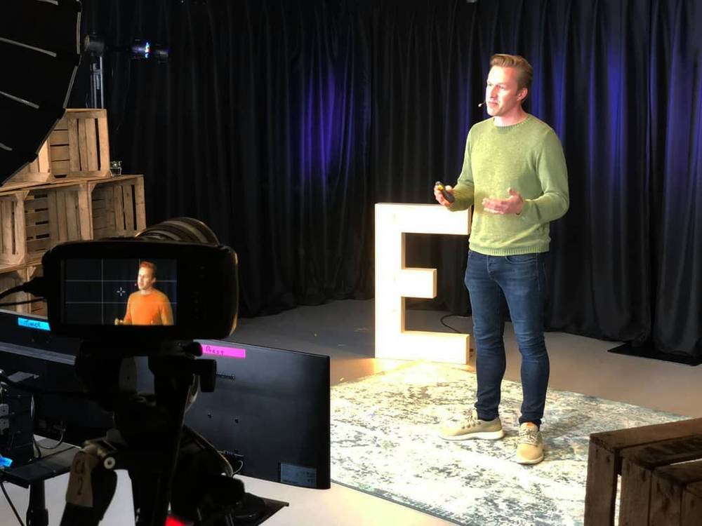 A man in a green sweater presenting on stage beside a lit letter E, with a camera monitor showing the live shot.
