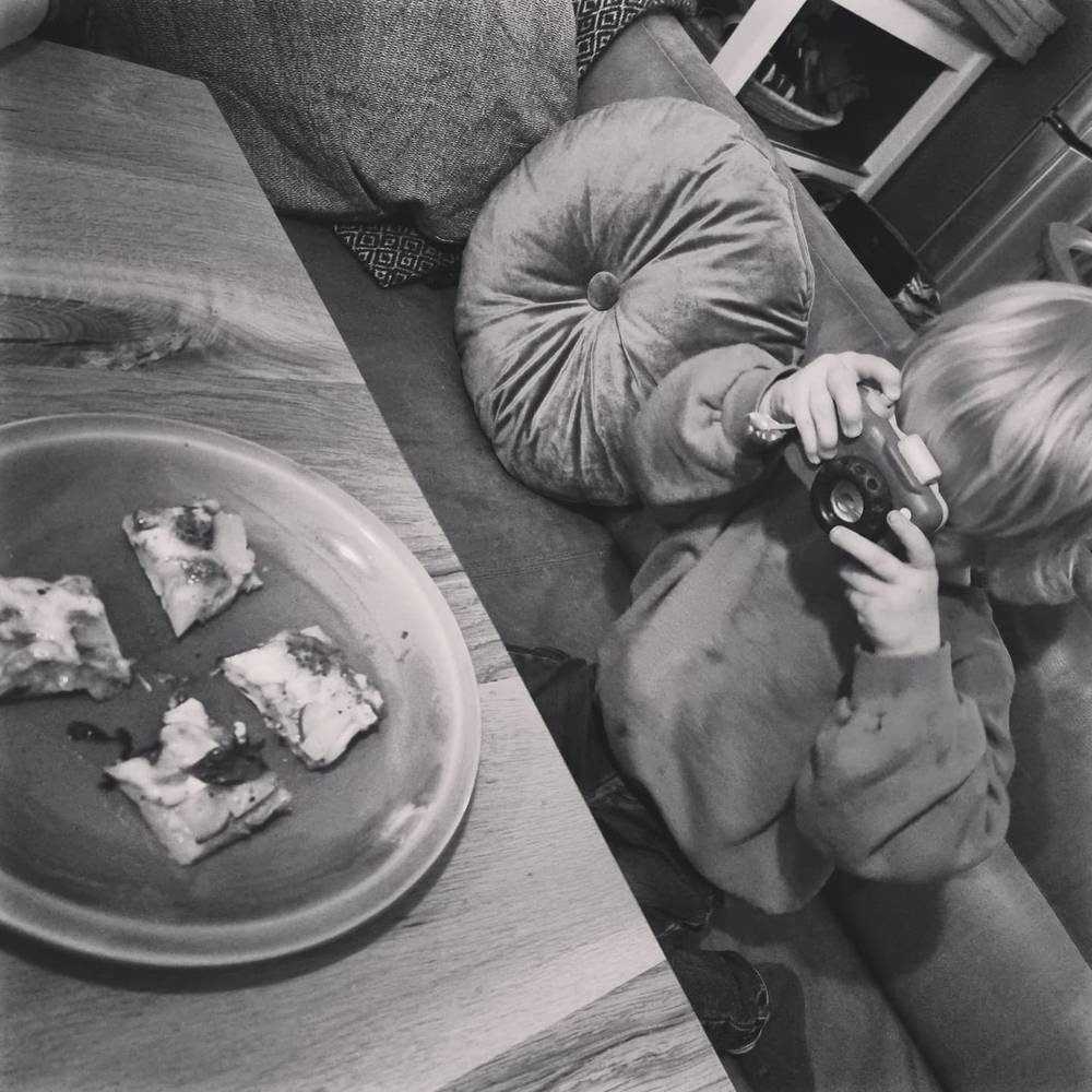 Black-and-white photo of a child holding a camera at the table, with small pizza slices on a plate nearby.