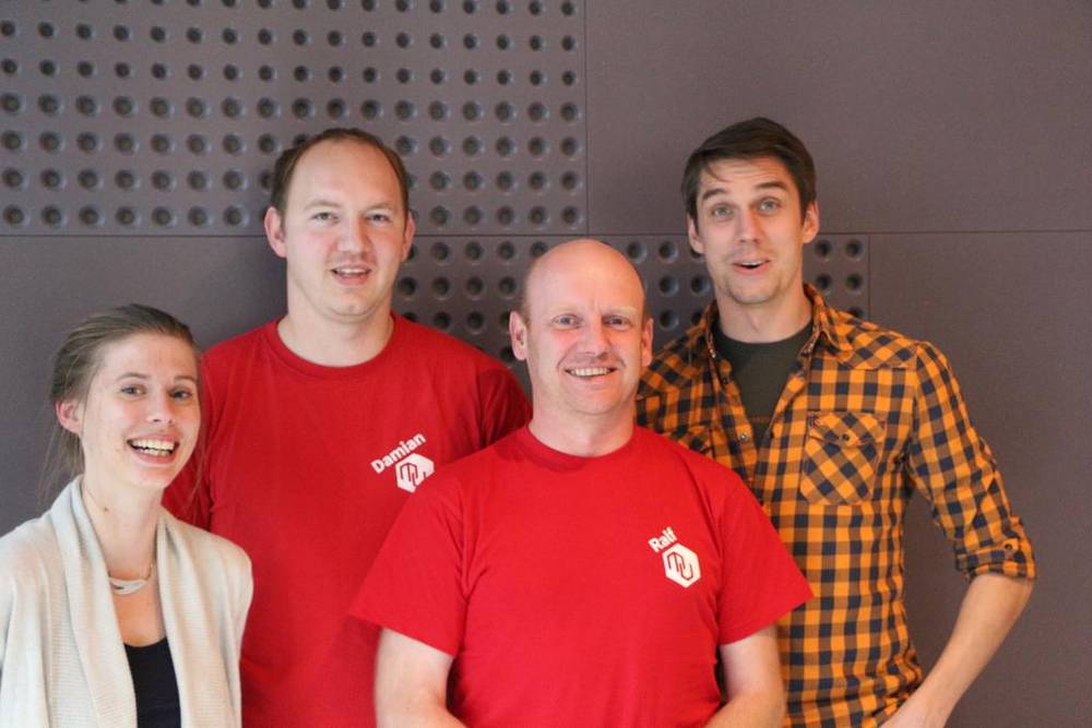 Four people, two wearing matching red t-shirts, posing together against a patterned wall.