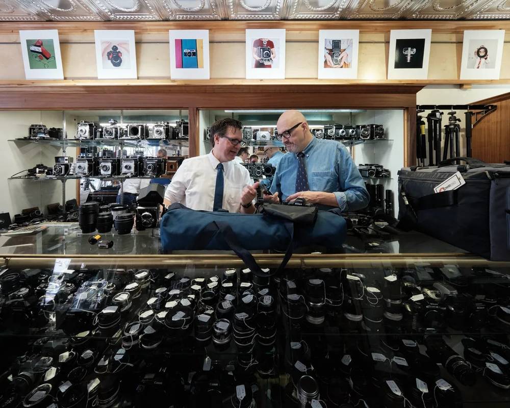 Mike Knight and Arthur Ruckle examine vintage cameras amid the organized chaos of Blue Moon Camera & Machine's Portland storefront. Warm tungsten light bathes the ornate tin ceiling and illuminates rows of meticulously arranged vintage cameras displayed on glass shelves, while hundreds of lenses carpet the floor below. The two specialists lean over their inspection table in focused collaboration, surrounded by decades of photographic history in this legendary Pearl District camera shop.