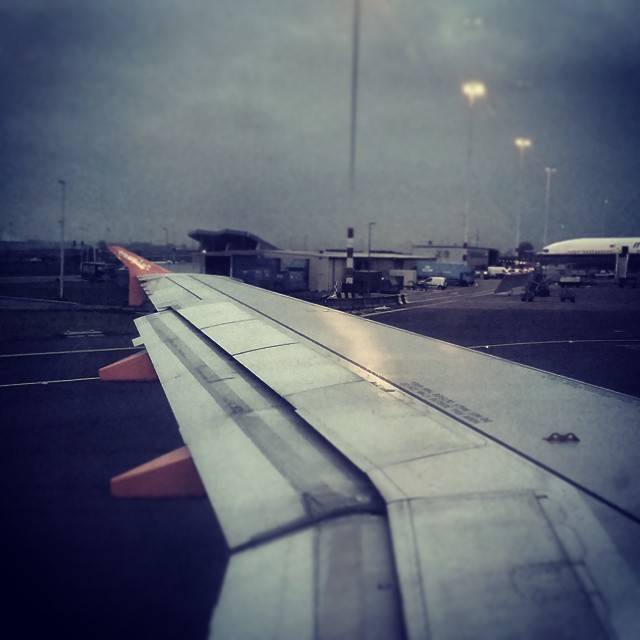 View of an airplane wing on the tarmac with airport buildings in the background.