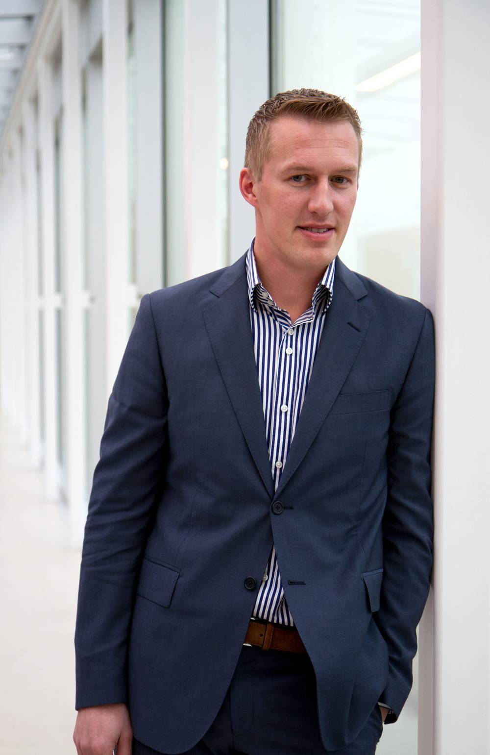 A portrait of a man in a blue suit standing against a bright hallway window.