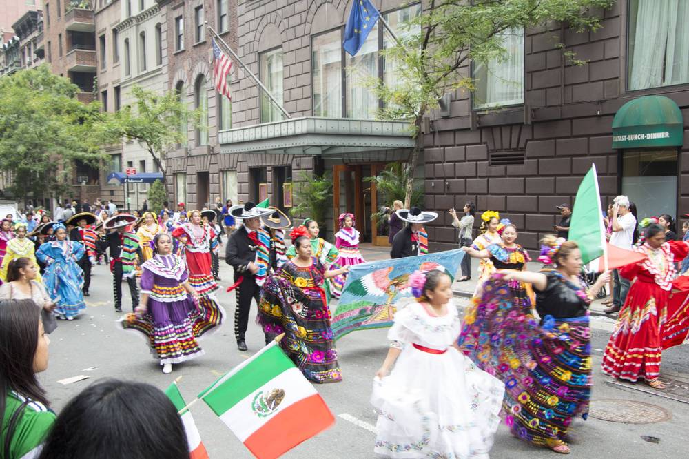 Folklórico dancers in colorful dresses parade down a Manhattan avenue waving Mexican flags.