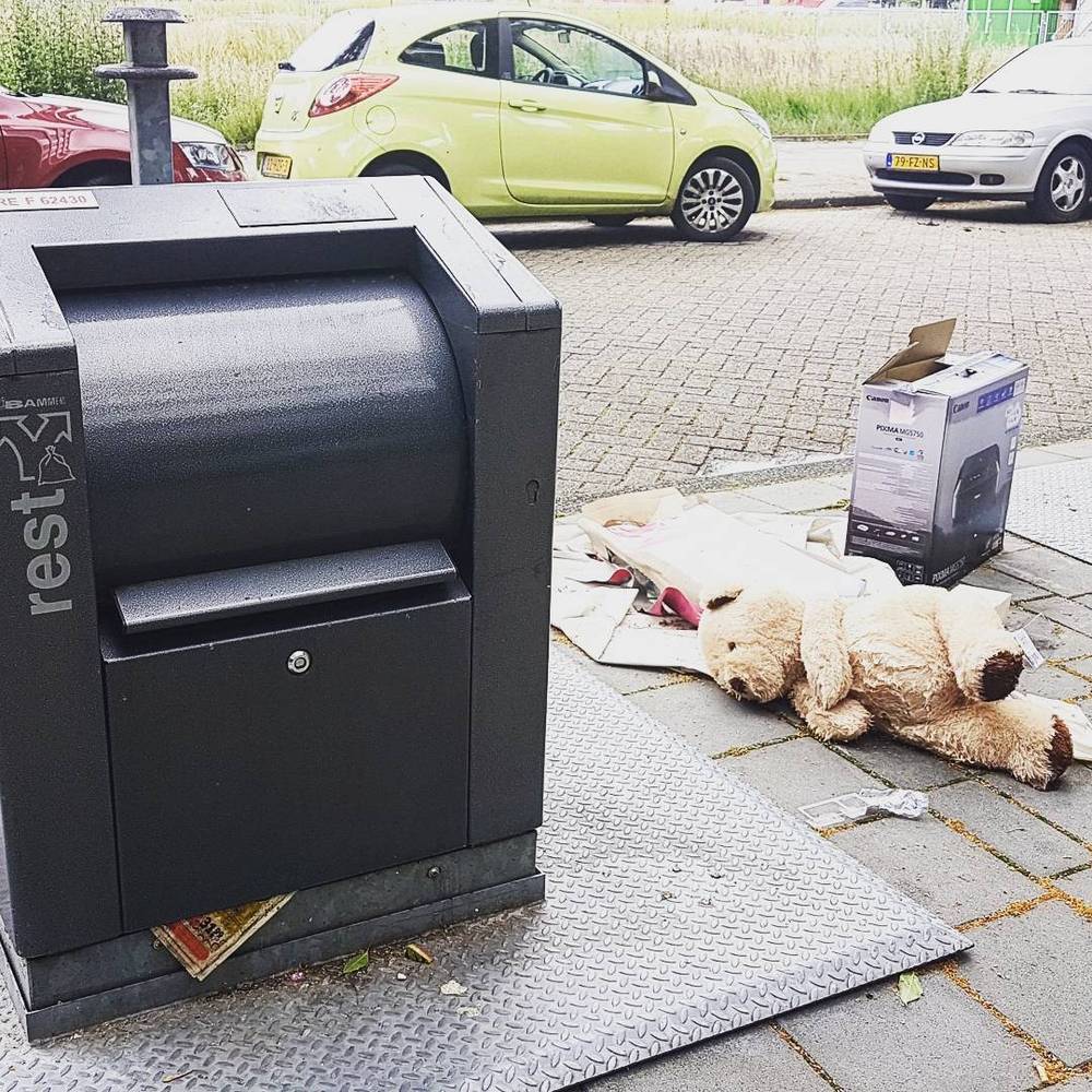 A rest household waste bin on a street beside a discarded stuffed toy and cardboard box.