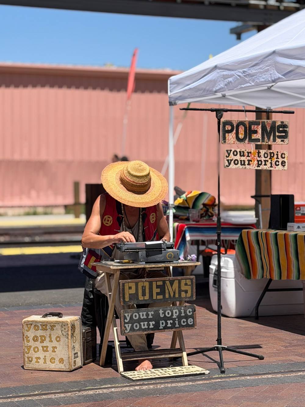 A man in a wide-brimmed hat sitting at a table in the sun, looking down at a typewriter. Signs surround him saying “Poems: your topic, your price”