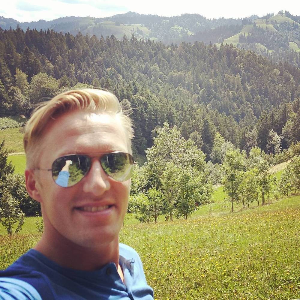 A selfie of a man in sunglasses with rolling green forested hills in the background.
