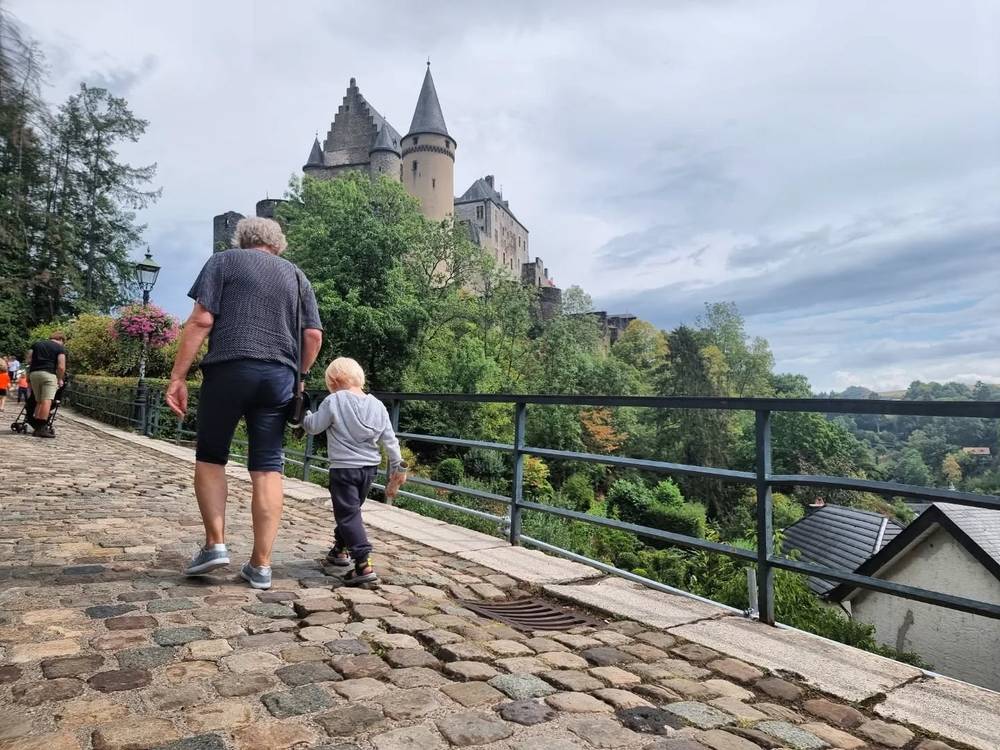 A man and a child walking together on a cobblestone path, with a hilltop castle visible in the distance.