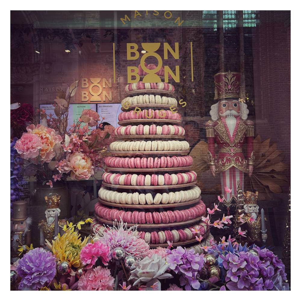 A window display for Maison Bon Bon in Bruges features a large, multi-tiered macaron tower stacked with alternating rows of pink and white macarons. The shop's gold logo is printed on the glass window directly above the tower.To the right stands a tall, ornate nutcracker soldier figurine dressed in a red and gold uniform with a tall hat. Two smaller nutcracker figurines sit near the bottom corners. The base of the display is filled with a dense, colorful arrangement of faux flowers in shades of pink, purple, and yellow.