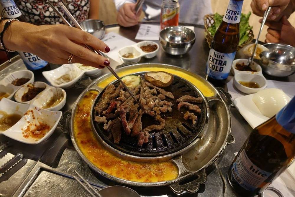 Meat grilling on a Korean barbecue tabletop grill, surrounded by side dishes and cold beer.
