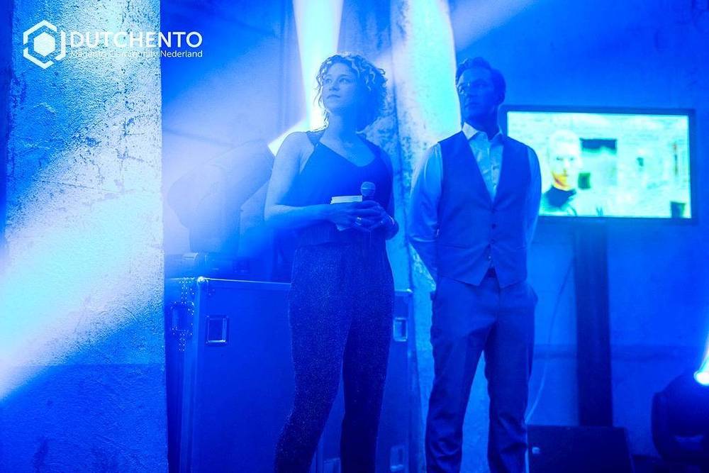 Two people standing on stage under blue lighting during a Dutchento event.