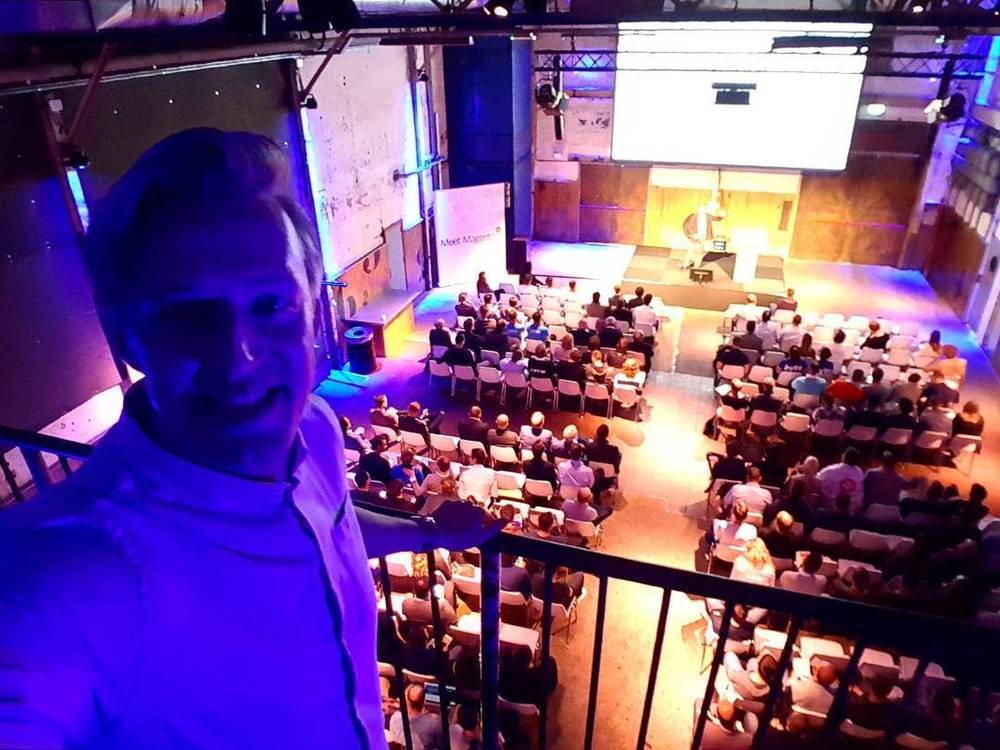 A man speaks to an audience from a balcony overlooking a lit conference stage below.