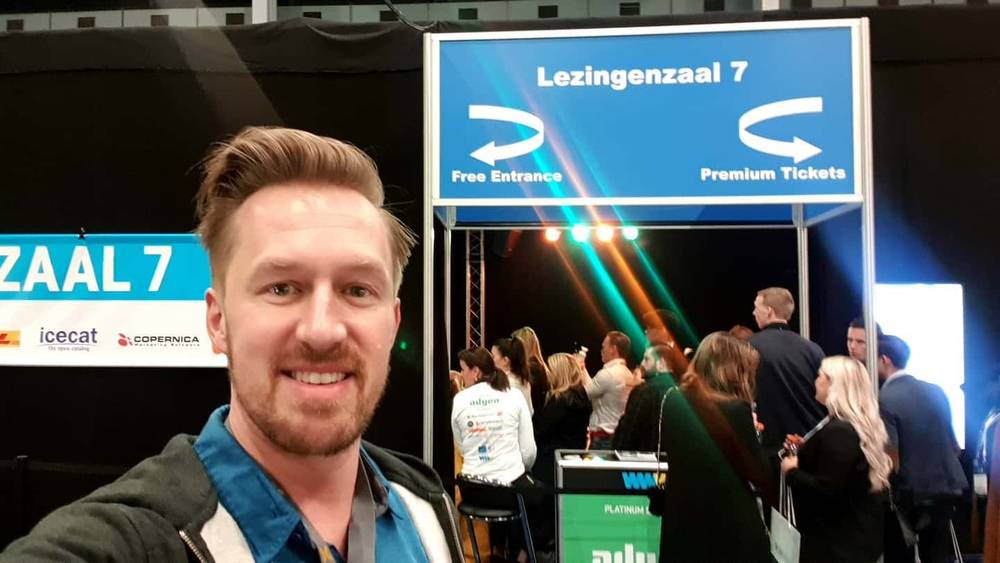 A man takes a selfie at a conference entrance sign for "Lezingenzaal 7" with people queuing behind him.