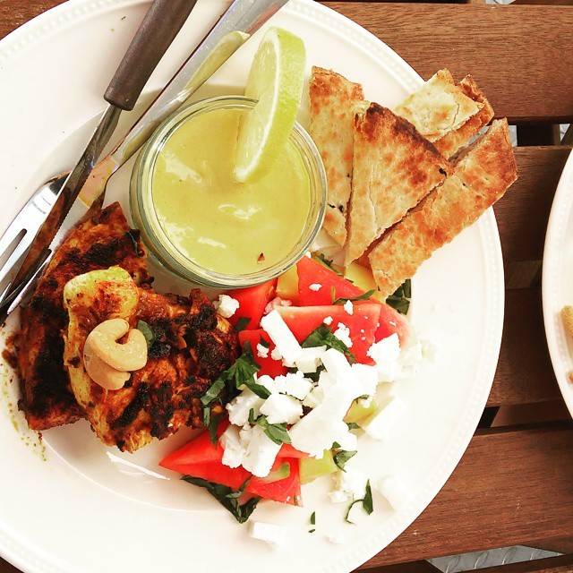 A plate of grilled meat, flatbread, tomato and feta with a small jar of dip garnished with lime.