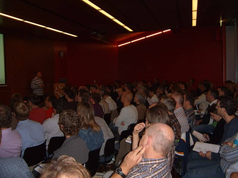 A large audience sits in rows in a dim conference hall watching a presentation.