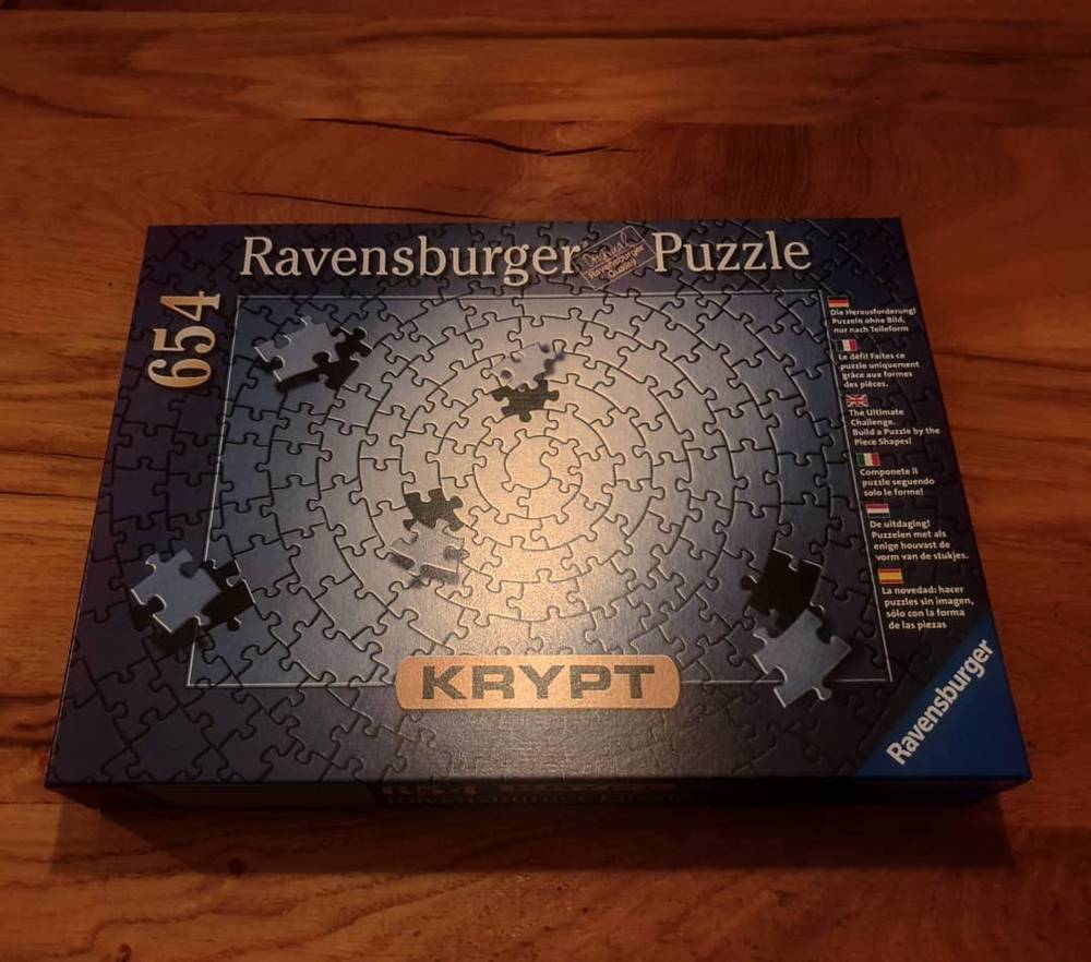 A box for a Ravensburger 654-piece Krypt jigsaw puzzle, entirely one color, sitting on a wooden table.