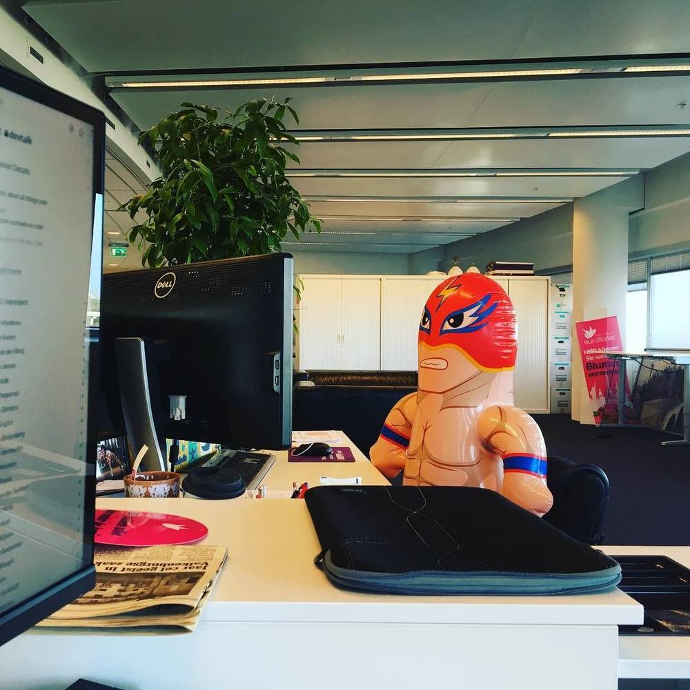 An inflatable wrestler figure sitting on an office desk in an open-plan workspace.