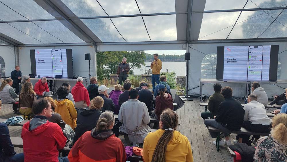An audience seated under a tent watches a presentation with screens showing app data.