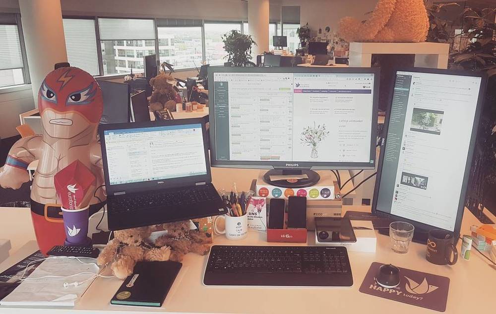 A cluttered desk with multiple monitors, a wrestler mascot figure, and office supplies.