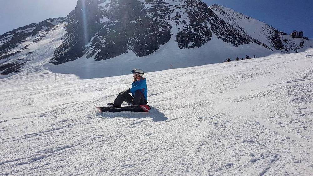 A lone snowboarder seated on a wide snow-covered slope near mountains.