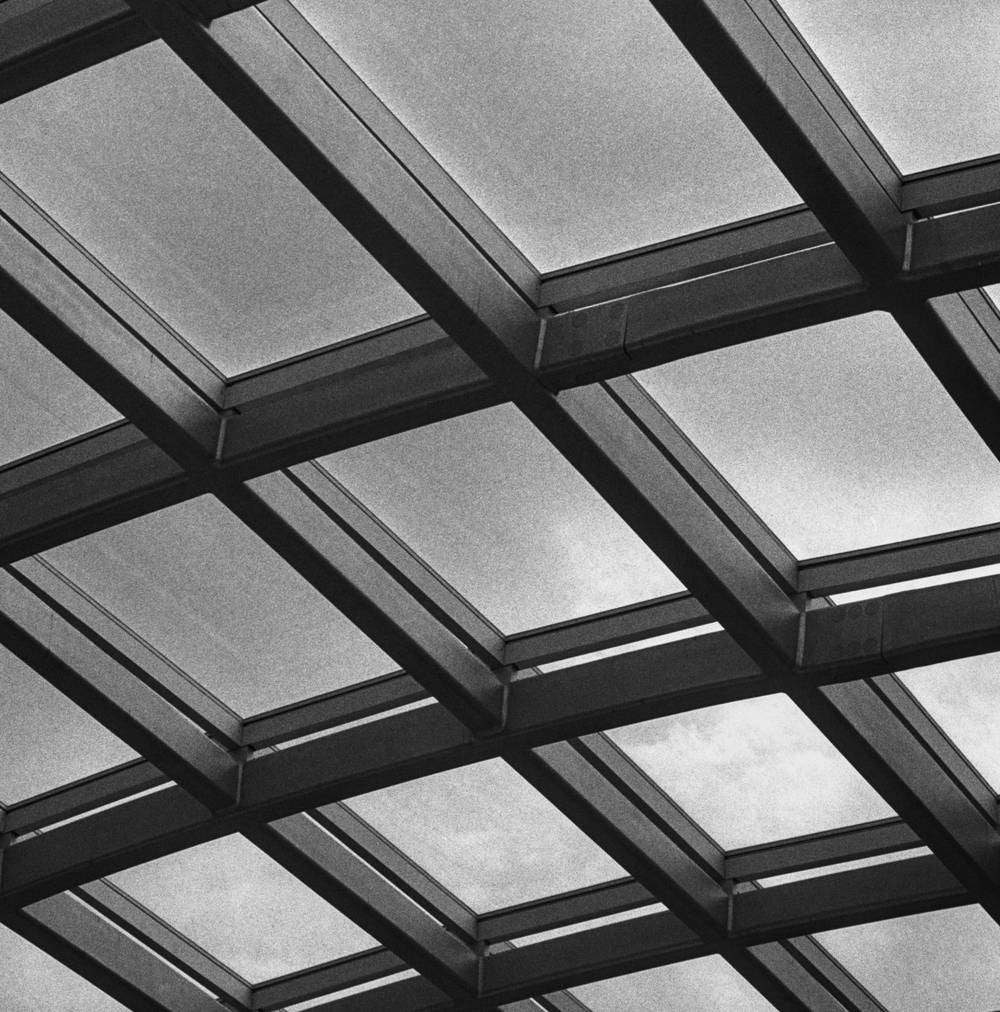 A black and white perspective view of a gridded glass skylight supported by a geometric framework of heavy metal beams.