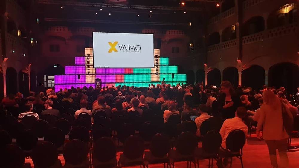The interior of an ornate historic venue with a stage screen showing the Vaimo logo, lit in red.
