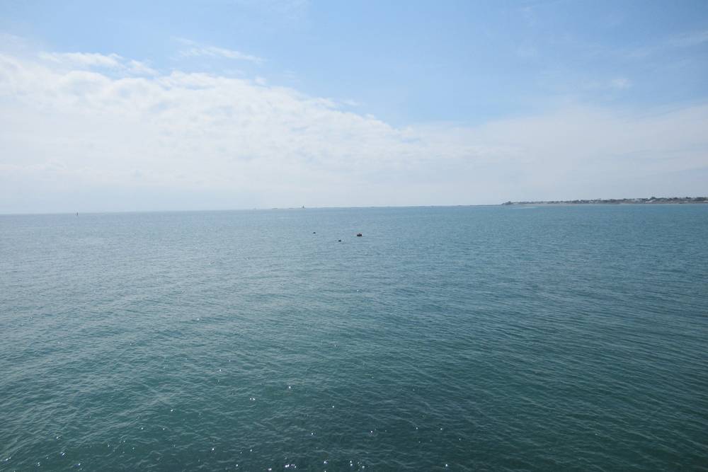A wide view of calm, turquoise sea water stretching to the horizon under a pale blue sky with light clouds. Small buoys bob in the distance, and a thin line of coastline is visible on the far right. The water's surface has gentle ripples and subtle sunlight glints.
