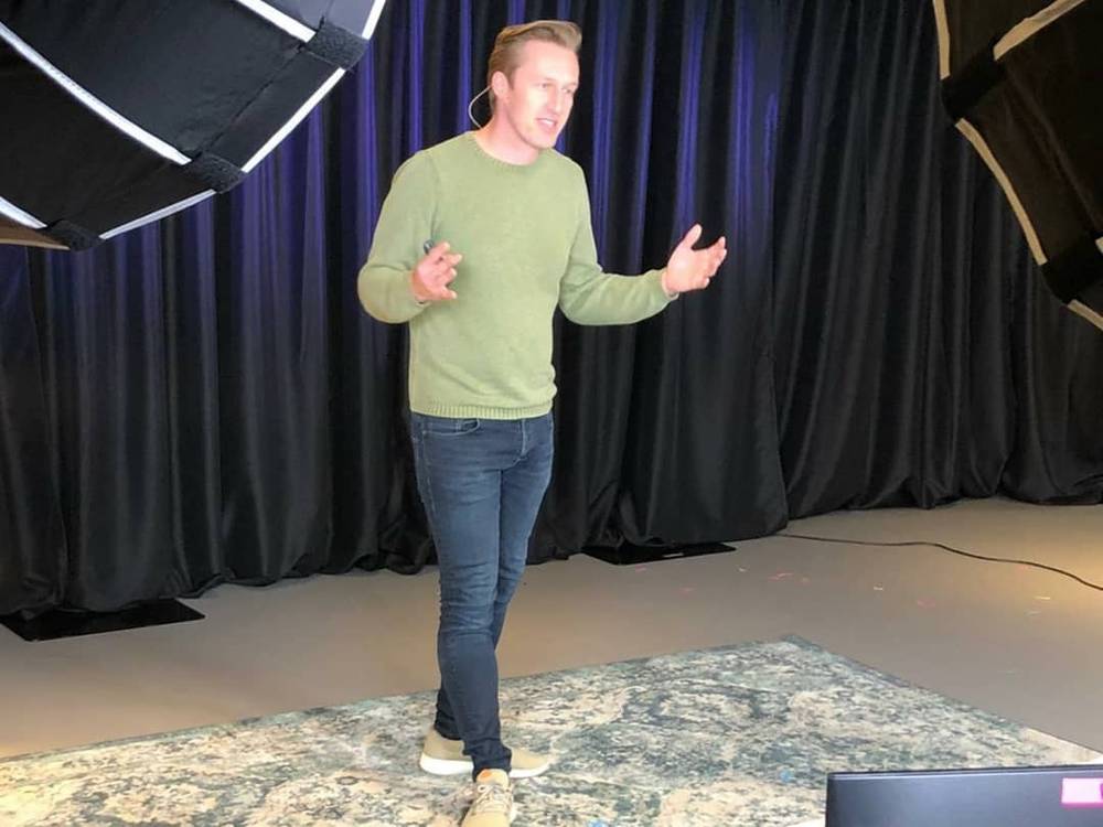 A man gesturing with both hands while presenting on a stage lit by studio lighting.