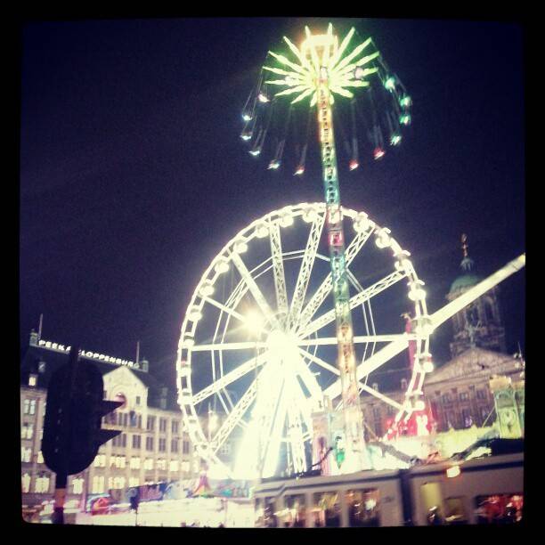 A lit-up ferris wheel and fairground ride at night in a town square.