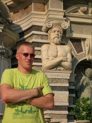 A man in a lime shirt poses with crossed arms beside an ornate stone statue.