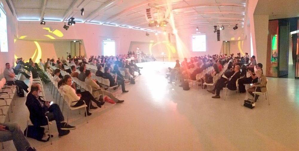 A packed conference audience hall with rows of chairs facing a stage at the far end.