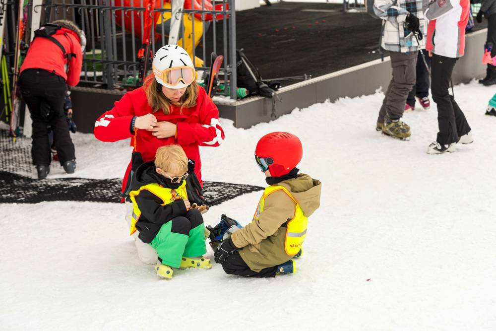 An adult in a red jacket helps two children in ski gear at the base of a slope.