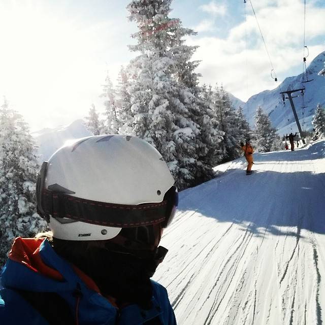 A skier in a white helmet is photographed close-up beside a sunny snow-covered slope with pine trees.