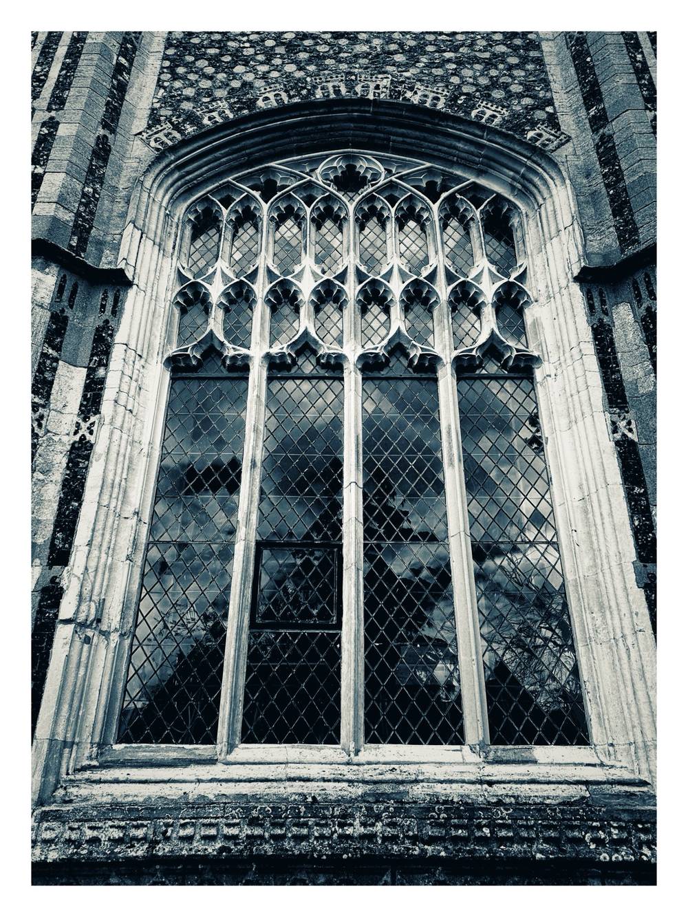 A black and white photograph captures a close-up, low-angle view of a large, ornate Gothic window set into a textured stone wall. The upper portion of the window features intricate, symmetrical stone tracery with delicate arches, while the main section consists of vertical panels divided by heavy stone mullions. Each panel is filled with a dense grid of diamond-shaped, leaded glass panes that reflect a dramatic sky with scattered clouds. The window is framed by deeply recessed stone molding, surrounded by flint masonry, and presented within a wide white border.