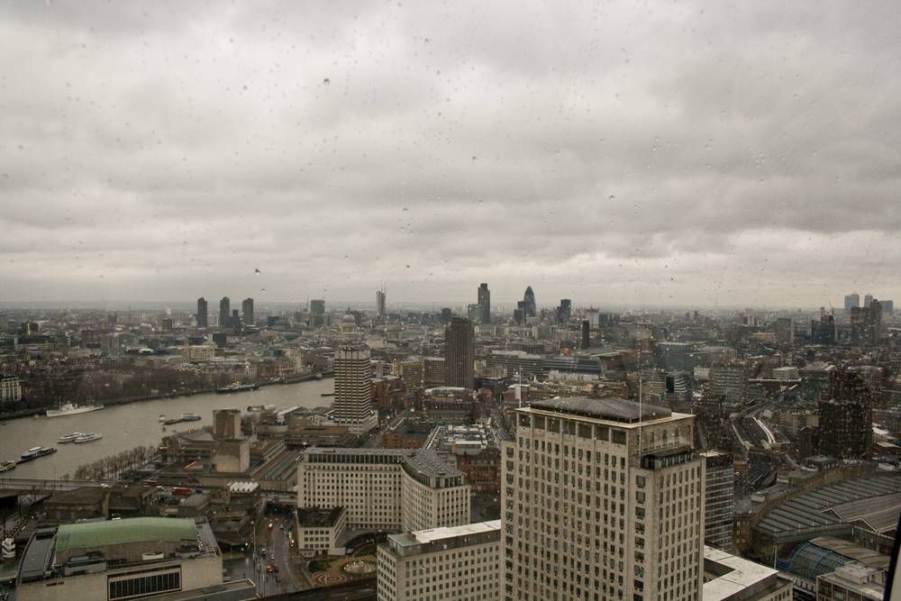 A hazy aerial view stretches over the London skyline and the Thames under an overcast sky.