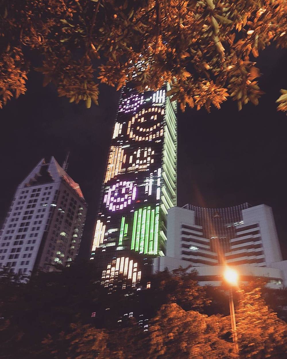 A skyscraper lit up with a colorful digital art display on its facade at night, framed by tree branches.