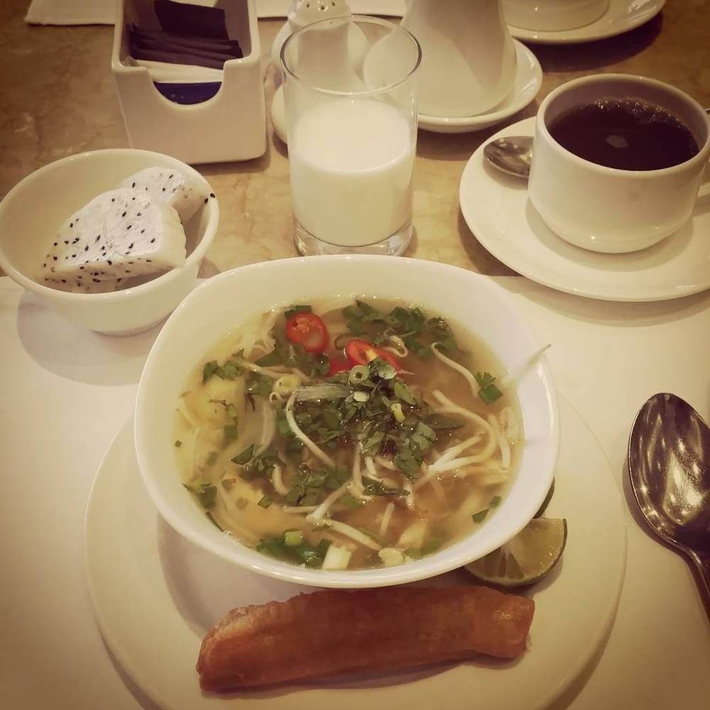 A bowl of pho soup on a table with a plate of fresh herbs, a glass of milk, and a cup of coffee.