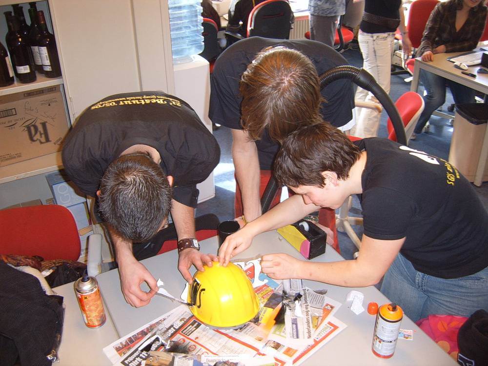 Three young men lean over a table painting and decorating a yellow safety helmet.