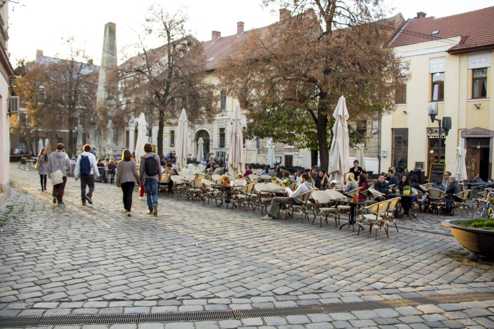 People walk and sit at outdoor cafe tables on a cobblestone square lined with trees.