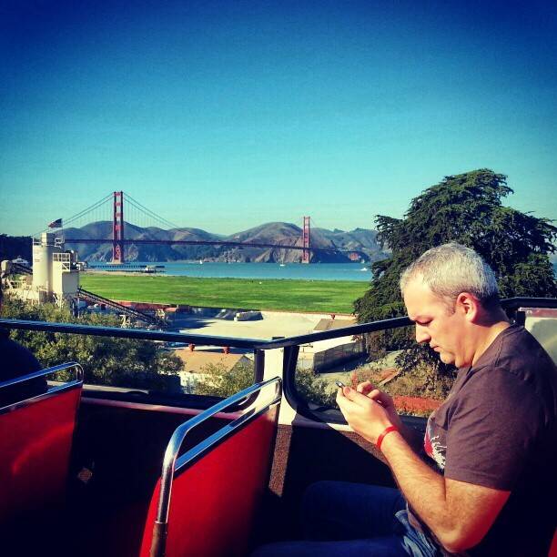 A man looks at his phone while riding an open-top tour bus with the Golden Gate Bridge visible behind him.