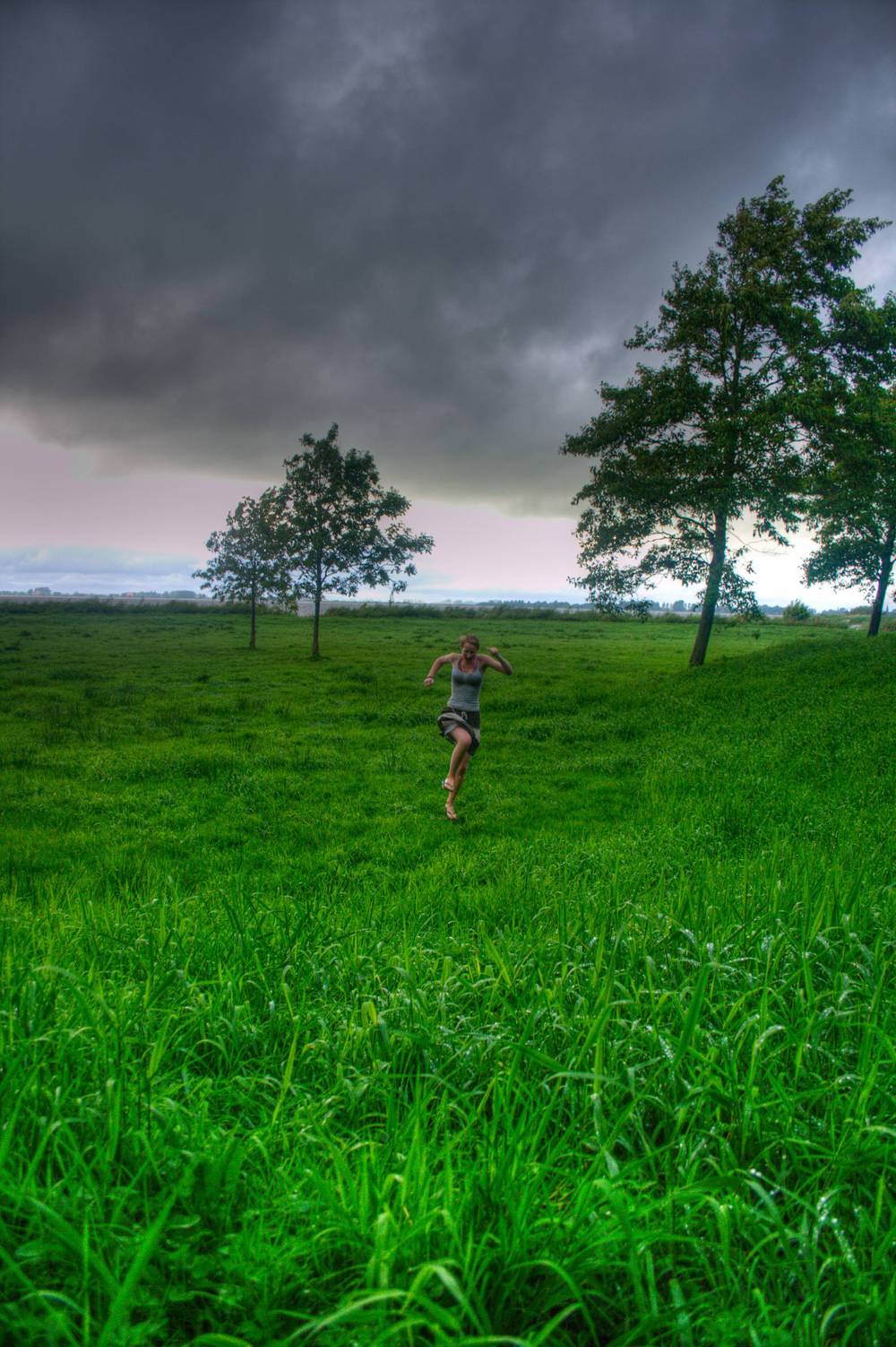 A woman runs across a grassy field beneath dark storm clouds and trees.
