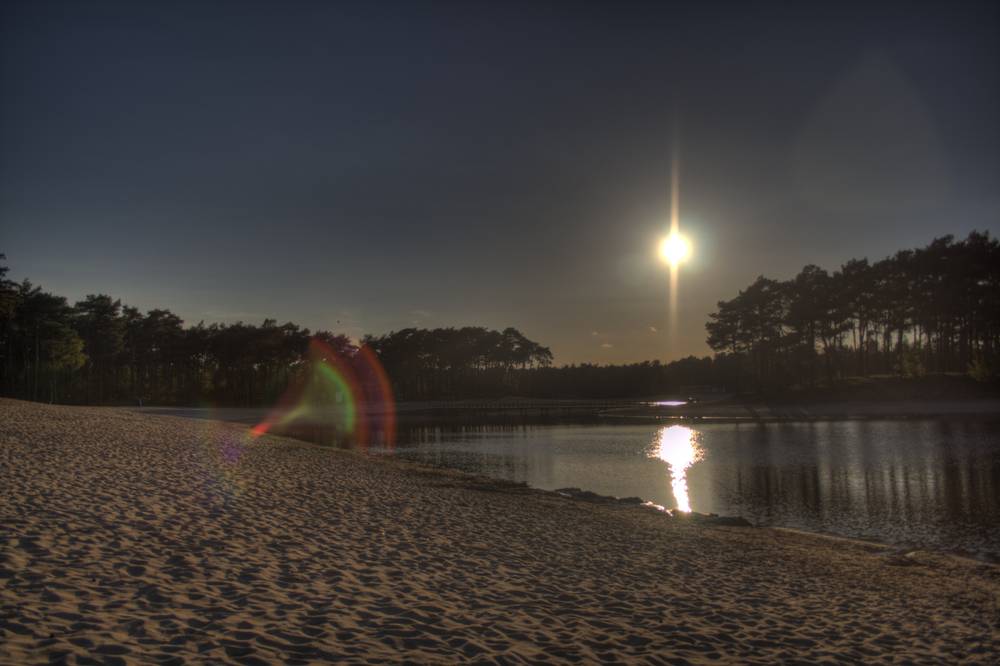 The sun sets behind trees at Henschotermeer lake, a lens flare glowing over the water.