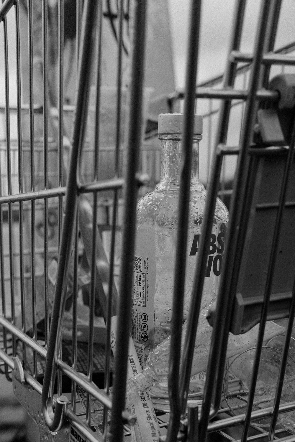 black & white photo, shot through the bars of a metallic shopping cart, focus on an empty vodka bottle and trash