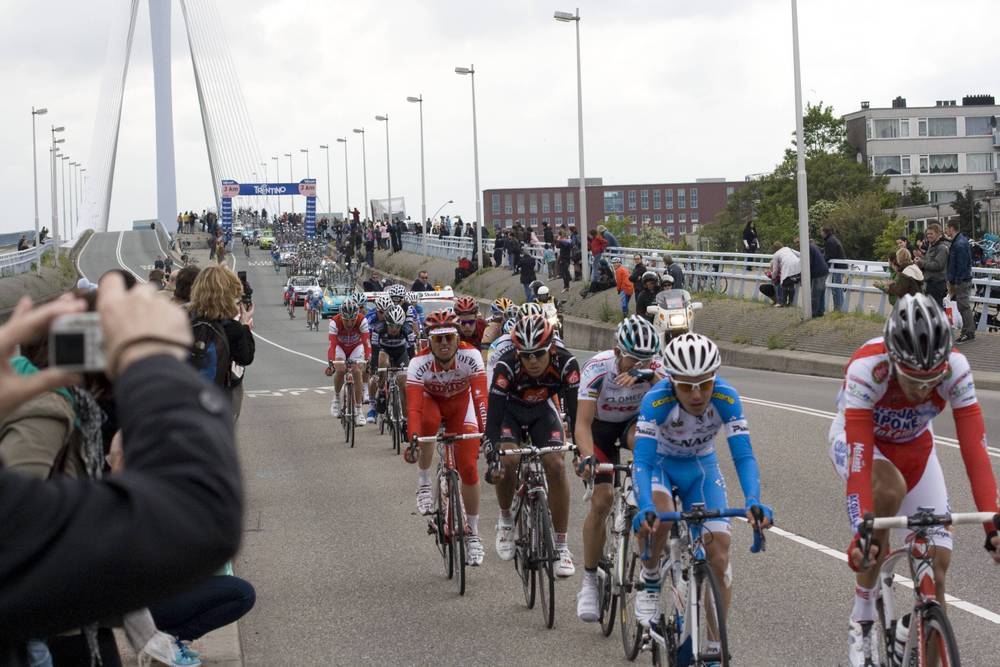 A pack of riders in red and white team kits sprints across the bridge, chased by photographers.