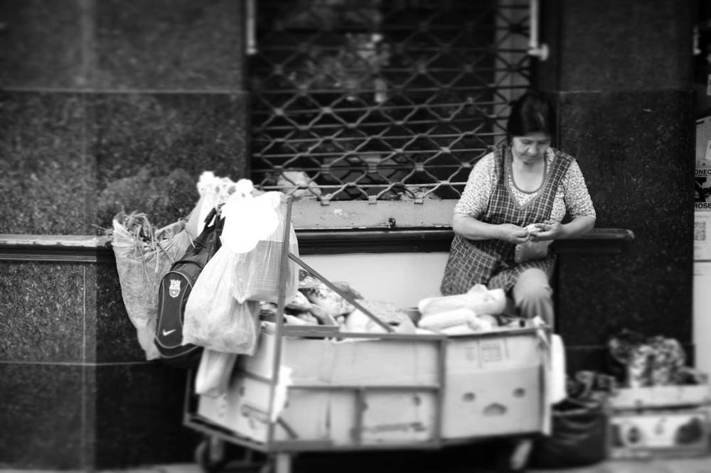 A black and white photo of a woman sorting items at a street cart loaded with bags.