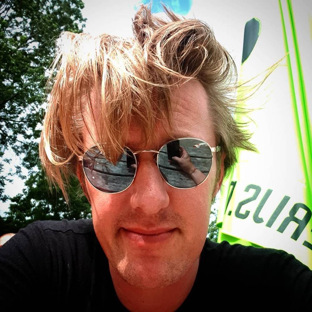 A close-up selfie showing windswept blond hair and sunglasses on a sunny, breezy day.