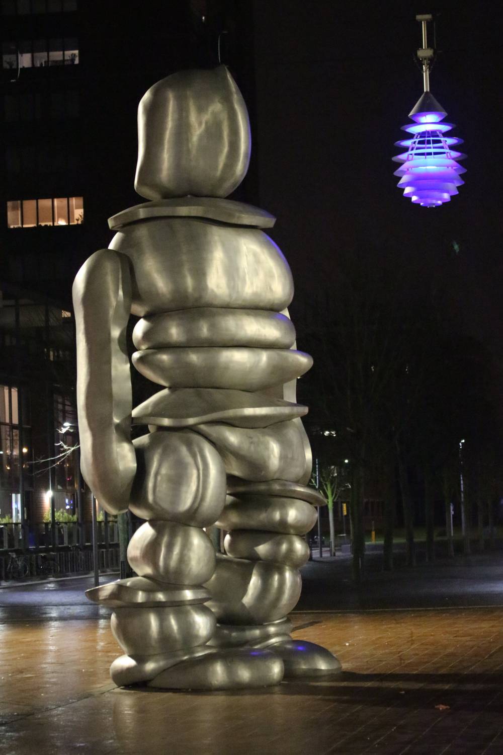 A silver sculpture of a hooded figure stands beside a glowing lamp in a plaza.