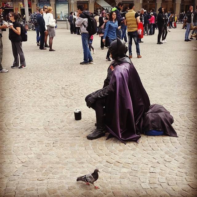 A street performer dressed as a caped, masked figure sits on the pavement in a busy square, with a pigeon nearby.