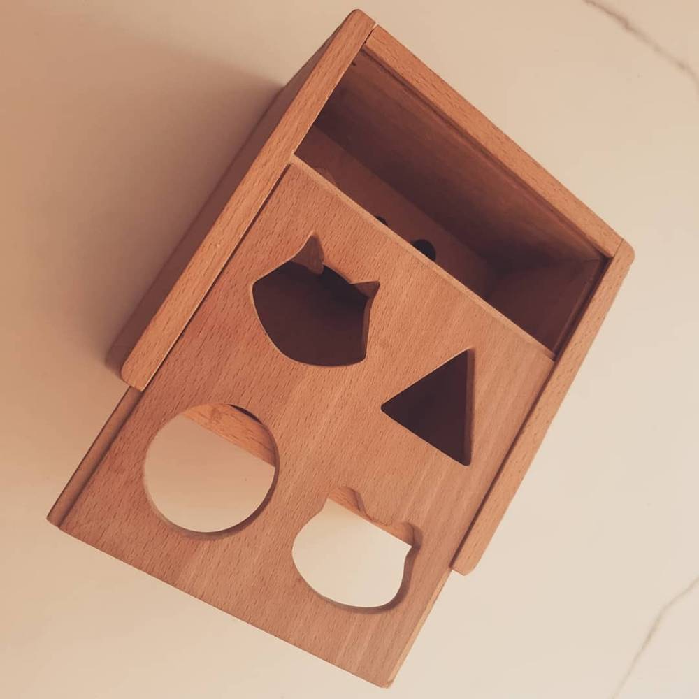 A wooden shape-sorting toy box with cut-out holes for a triangle, circle, square, and other shapes.