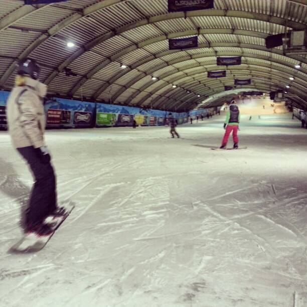 Two people ski and snowboard down an indoor artificial snow slope under an arched roof.